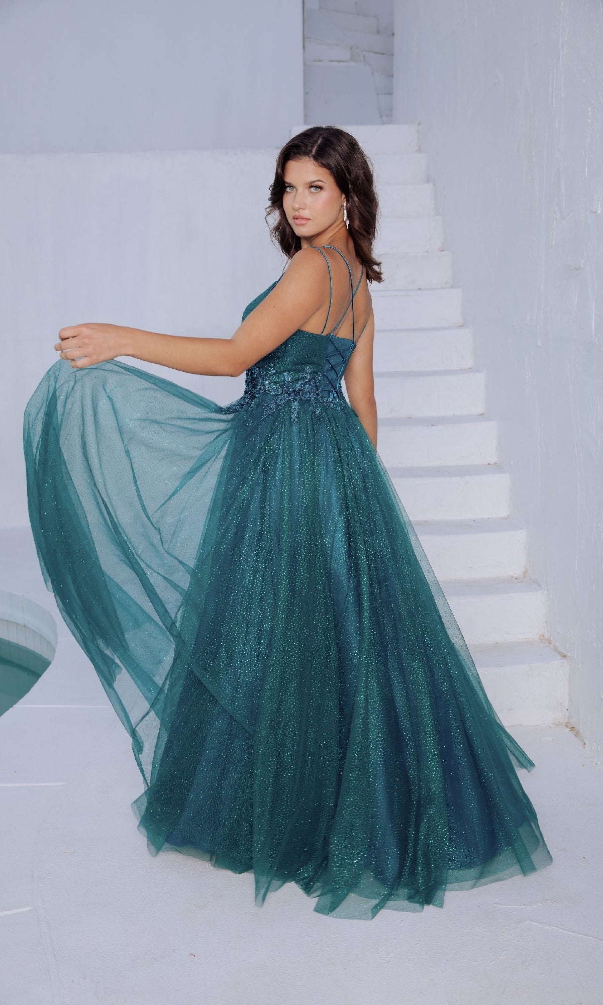 A woman in the Eureka 9797 Lace-Up Long Shimmer Prom Dress with thin straps stands by white stairs, holding out part of her green gown and looking over her shoulder.